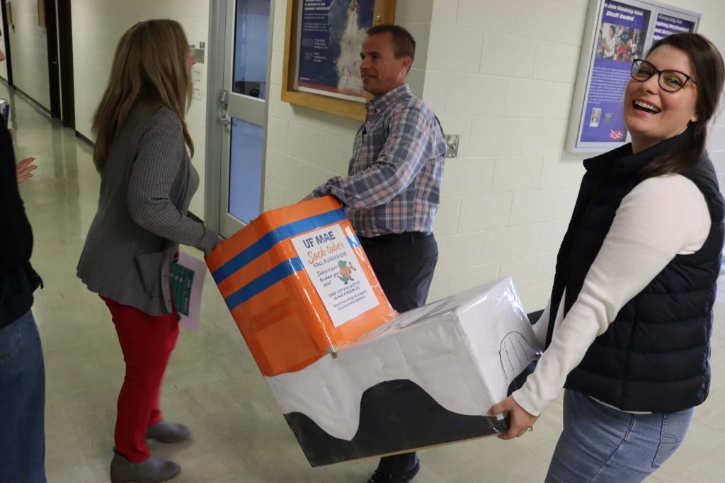 MAE team members carry sock shaped box down a hallway while smiling.