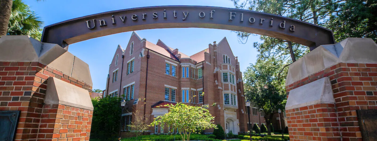 Entrance arch reading “University of Florida” framing a red brick campus building beneath a clear blue sky.