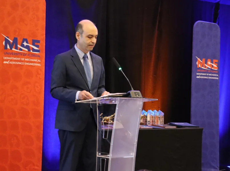 Pedro Mago standing at a Podium with orange and blue banners behind him.